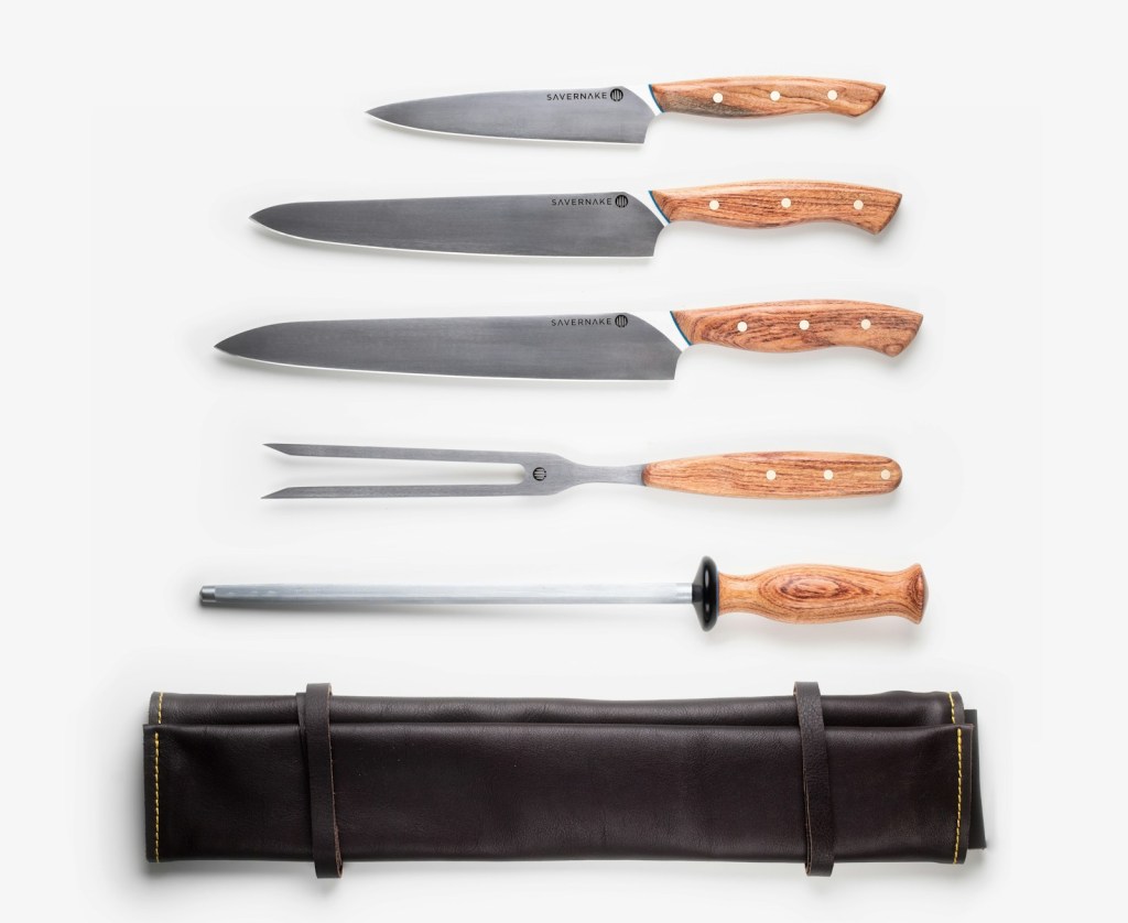 Three chef's knives, a carving fork, and a honing steel with wooden handles laid out above a rolled leather knife roll